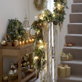 Lit candles on wooden hallway table next to staircase decorated with golden ribbons, branches and pine cones next and gifts on the stairs