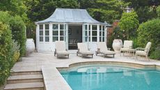 A traditional garden with paved patio and pool, sun loungers and chairs, and a white pool house. Lush hedges surround the space.