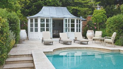 A traditional garden with paved patio and pool, sun loungers and chairs, and a white pool house. Lush hedges surround the space.