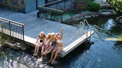 Four smiling women sit on a dock and wave.