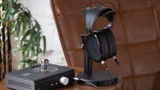 Dan Clark Audio Noire XO open-back headphones on a stand next to a headphone amp. There is a brown leather chair and a pot plant in the background