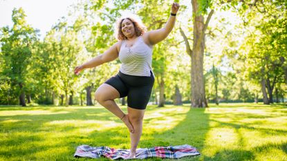 A woman stands in a leafy park balancing on one leg on a blanket.