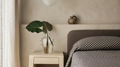 bedroom with plaster walls with niche behind the bed, side table with glass vase with branches, and gray headboard with checkered bedcover