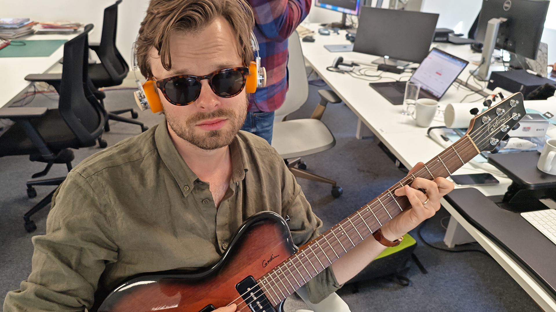 A man wearing orange headphones, plays a brown guitar, in an office.