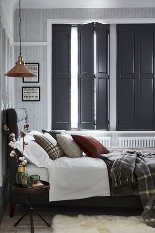 grey and white bedroom with grey textured wallpaper, dark grey shutters, white bedlinen, grey check blanket, wooden floor, copper pendant