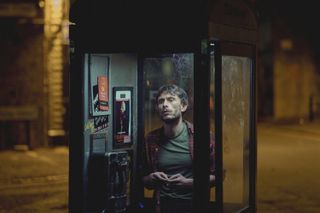 Richard Gadd as Donny, standing in a glass-walled phone booth at night, in the netflix show 'baby reindeer'