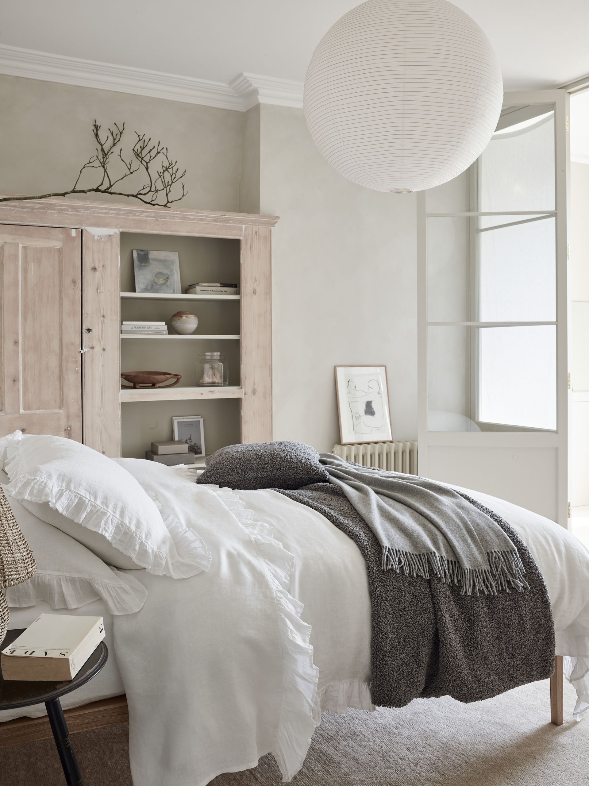 white and neutral bedroom with limed cabinet, stone walls, white bedding, cream carpet, gray blanket and cushion, white paper lampshade