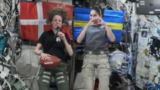 two astronauts floating in a module. one holds a football. flags are behind them