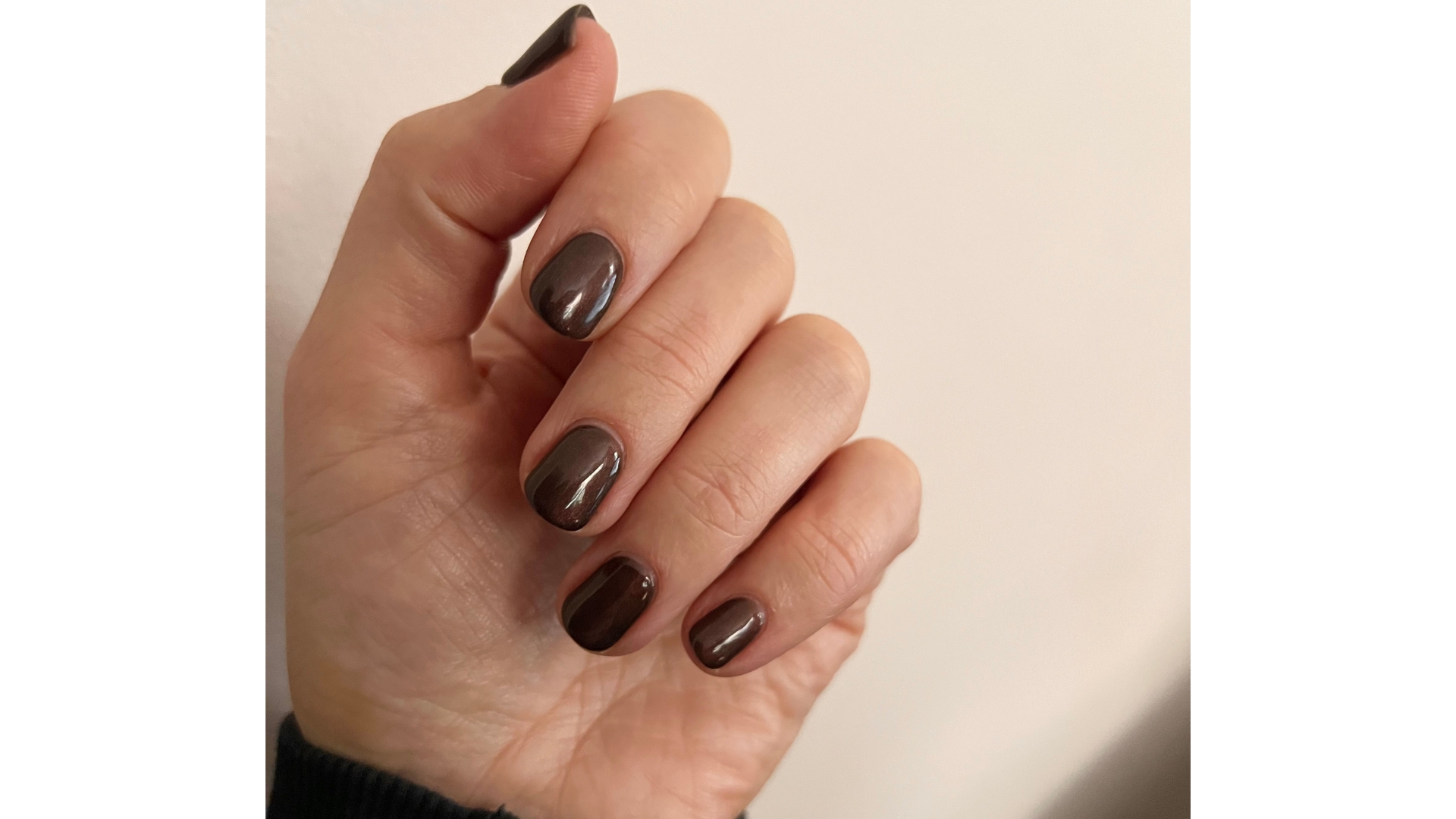 A close-up of Digital Beauty Editor, Fiona McKim&#039;s hand, showcasing her short and shimmery dark brown, cola-inspired manicure/ pictured in front of a plain cream background