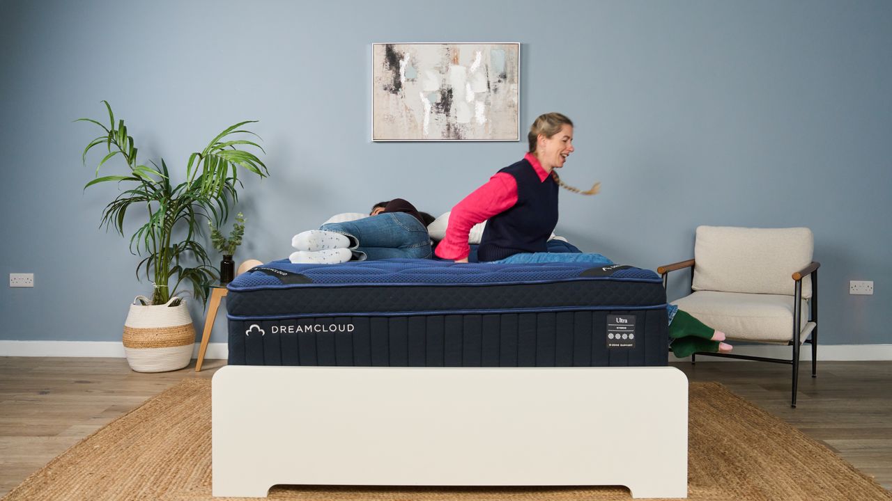 The image shows a mattress tester lowering herself onto a navy blue mattress while another tester lies on it on her side