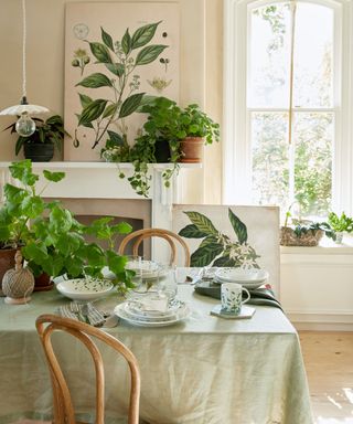 A white dining area with large windows, a white decorative fireplace, a mint green dining table flanked by wooden loop chairs, white dishes, and plants on and around the table. ing