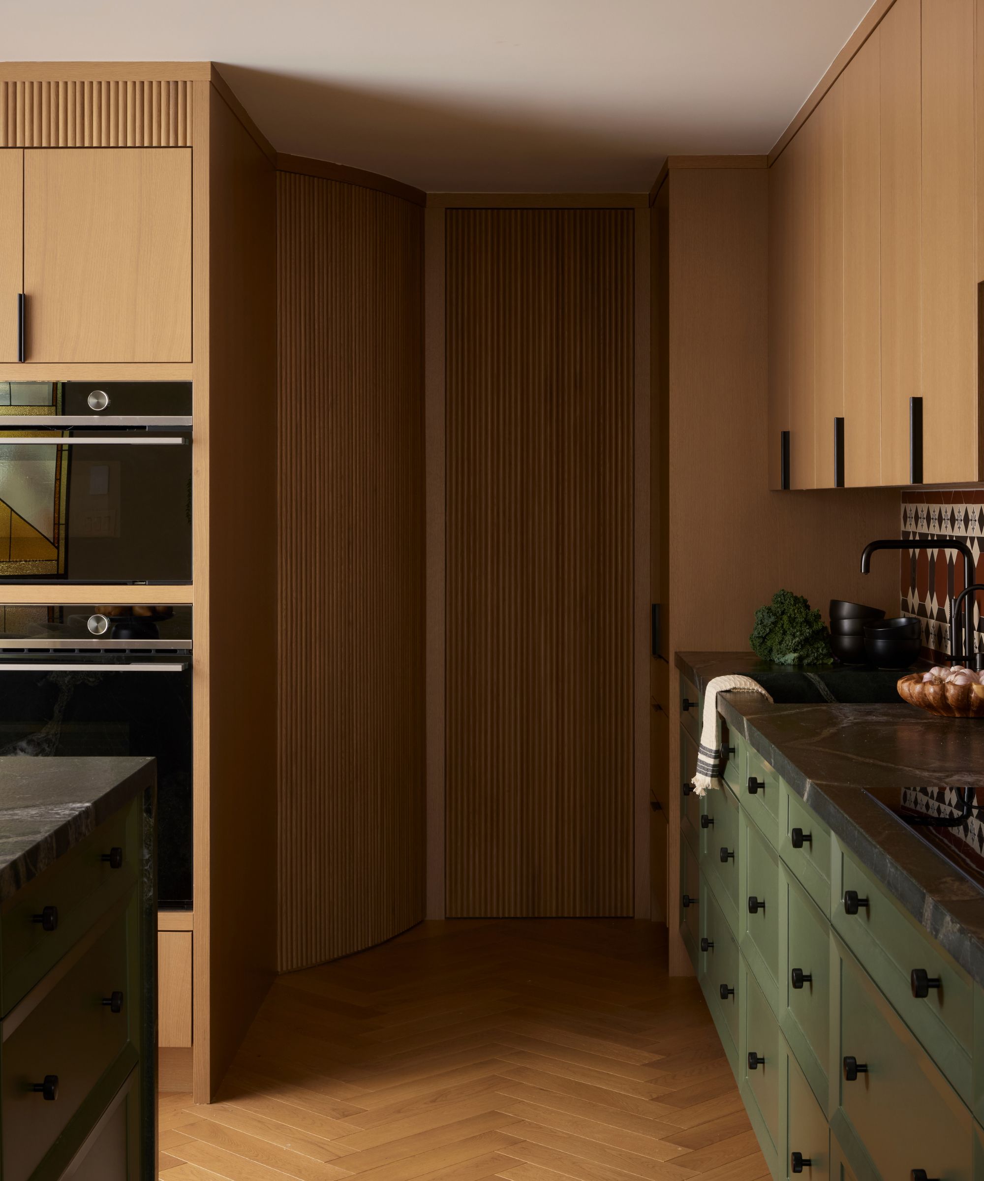 Moody kitchen with dark green lower cabinets, black hardware, a dark marble countertop, and a curved, vertically slatted wood accent wall in a herringbone hallway.