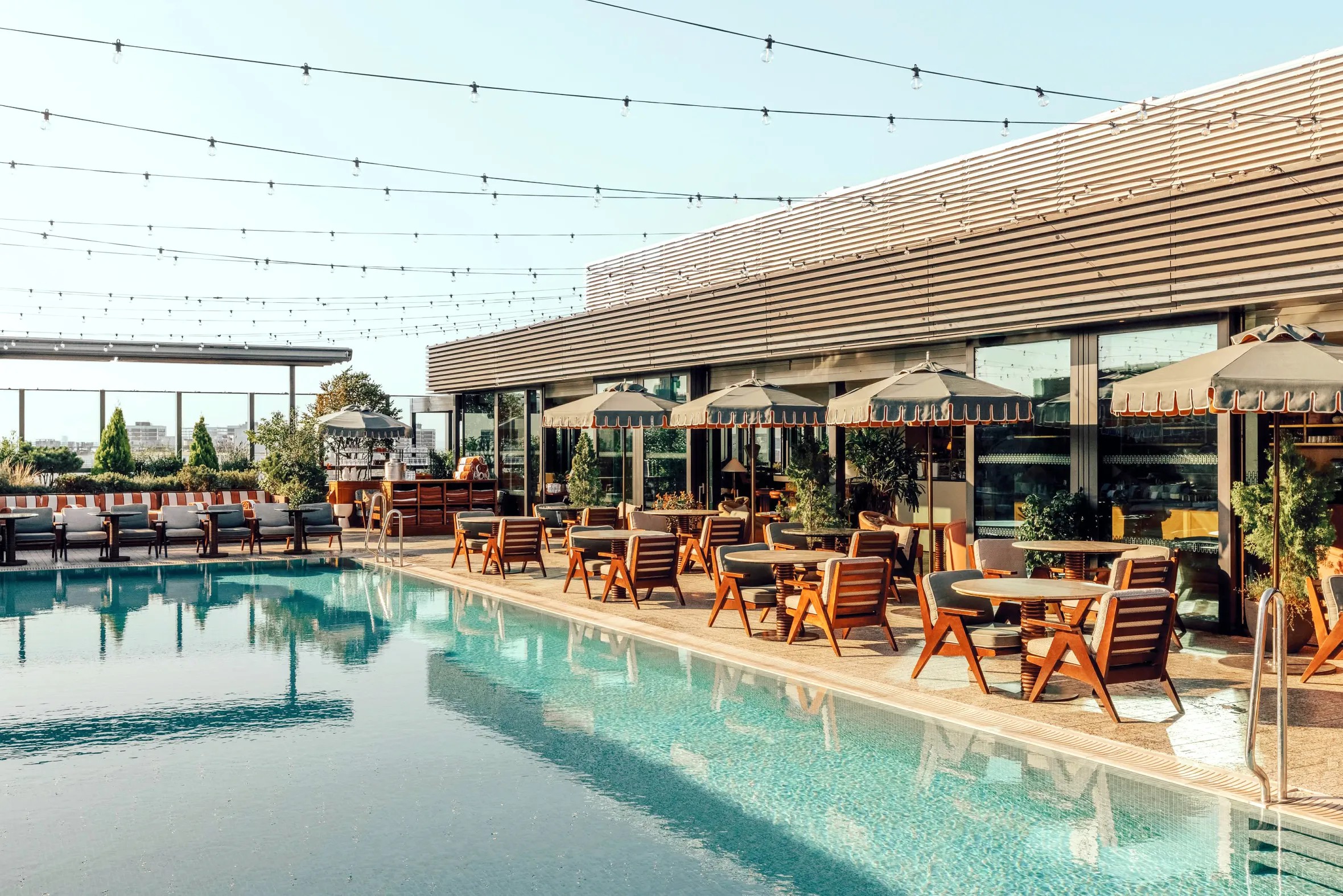 A sun-lit rooftop terrace with wood and textile loungers and dining tables, blue and red parasols, and lines of fairylights floating over it.