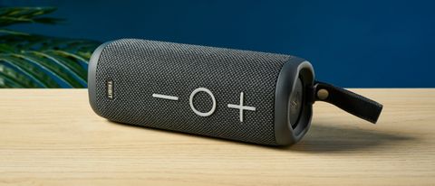 a black oblong-shaped bluetooth speaker with mesh covering and a carry strap photographed on a beige table in front of a navy wall