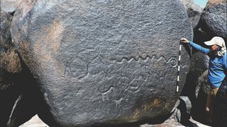 A person stands measures rock art depicting a snake. 