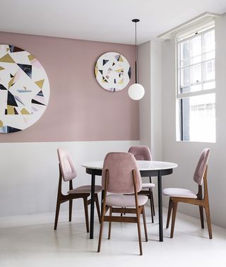 A round white table with 4 pink and white chairs in a corner next to a pink wall with round decorations on the wall and a window on the other side of the table