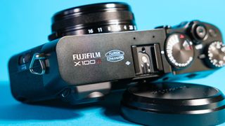 The Fujifilm X100VI mirrorless camera against a blue background lying on its side propped up using the lens cap.