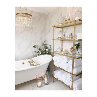 Glam bathroom with freestanding bathtub and open shelving unit in brass and glass styled with rolled white towels, toiletries and decorative accessories.