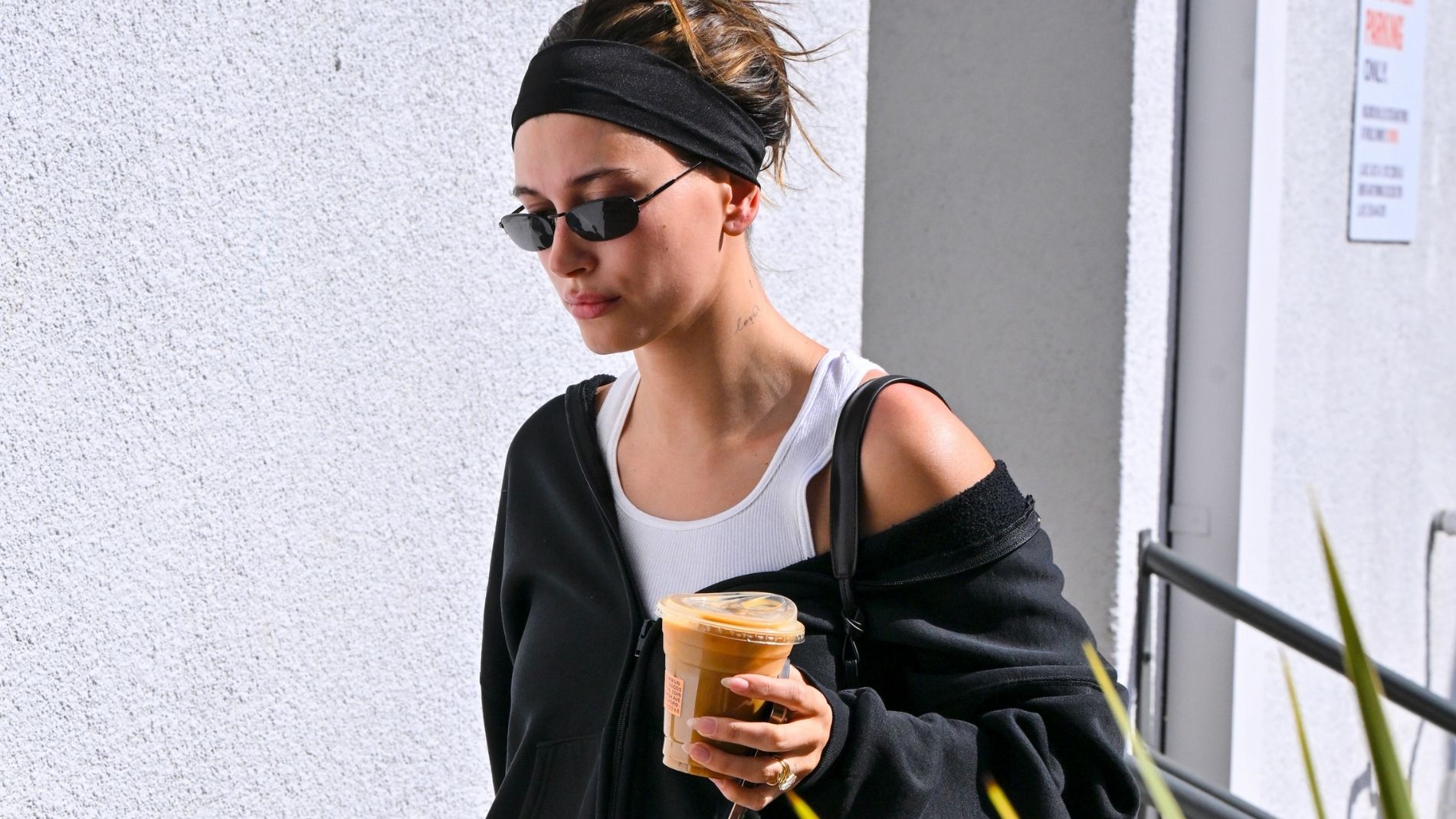 Hailey Bieber wears a black exercise outfit with sunglasses and a headband while carrying a cup of ice coffee. 