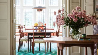 A wooden dining table and chairs visible through an open doorway from an entryway. A large wooden table with pink flowers in a vase in the foreground.