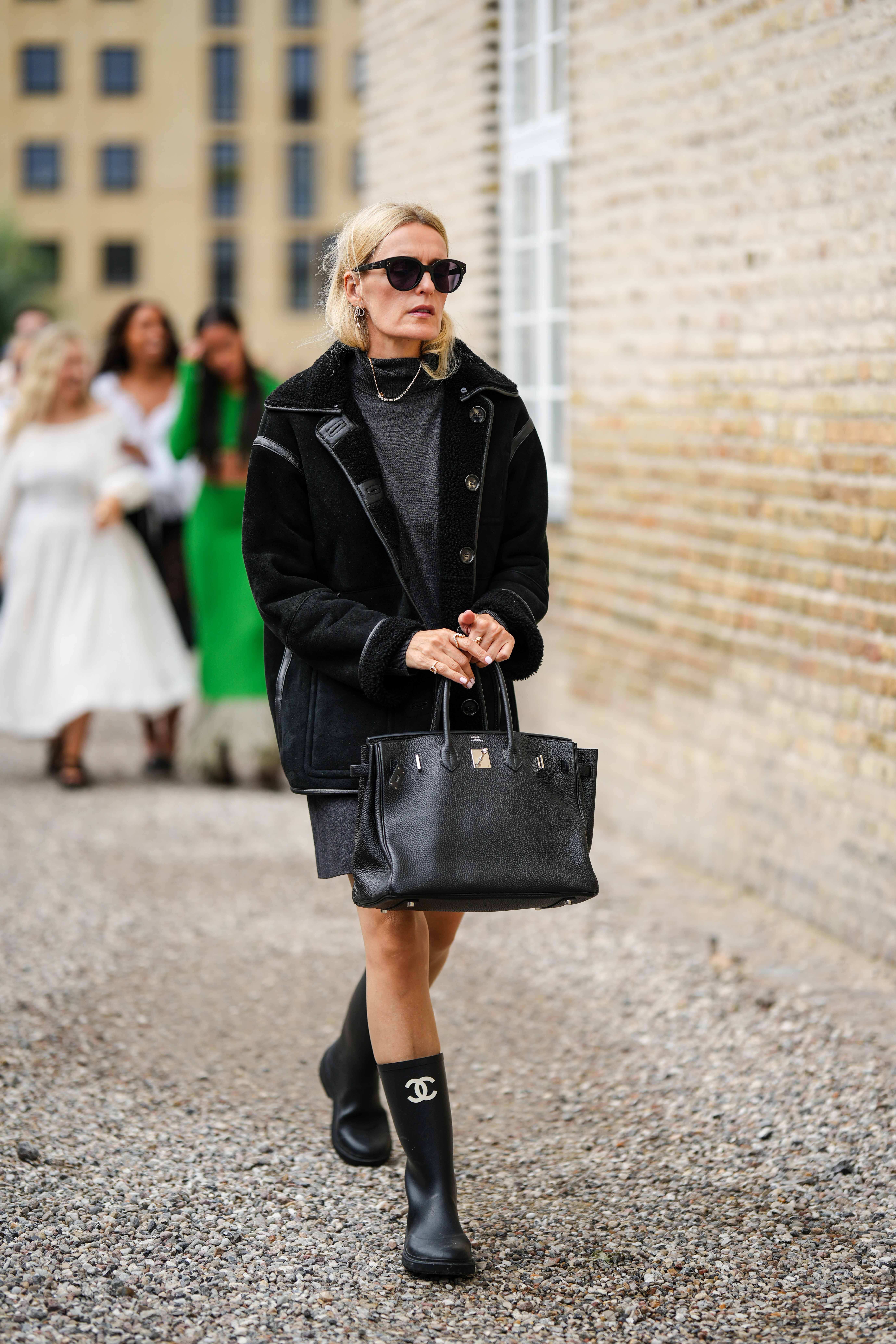 A guest wears black sunglasses, a dark gray wool short t-shirt dress, a black sheep and suede long jacket, a black shiny leather Birkin handbag from Hermes, a gold necklace, a black shiny plastic ankle boots from Chanel , outside Munthe, during the Copenhagen Fashion Week Spring/Summer 2024 on August 10, 2023 in Copenhagen, Denmark