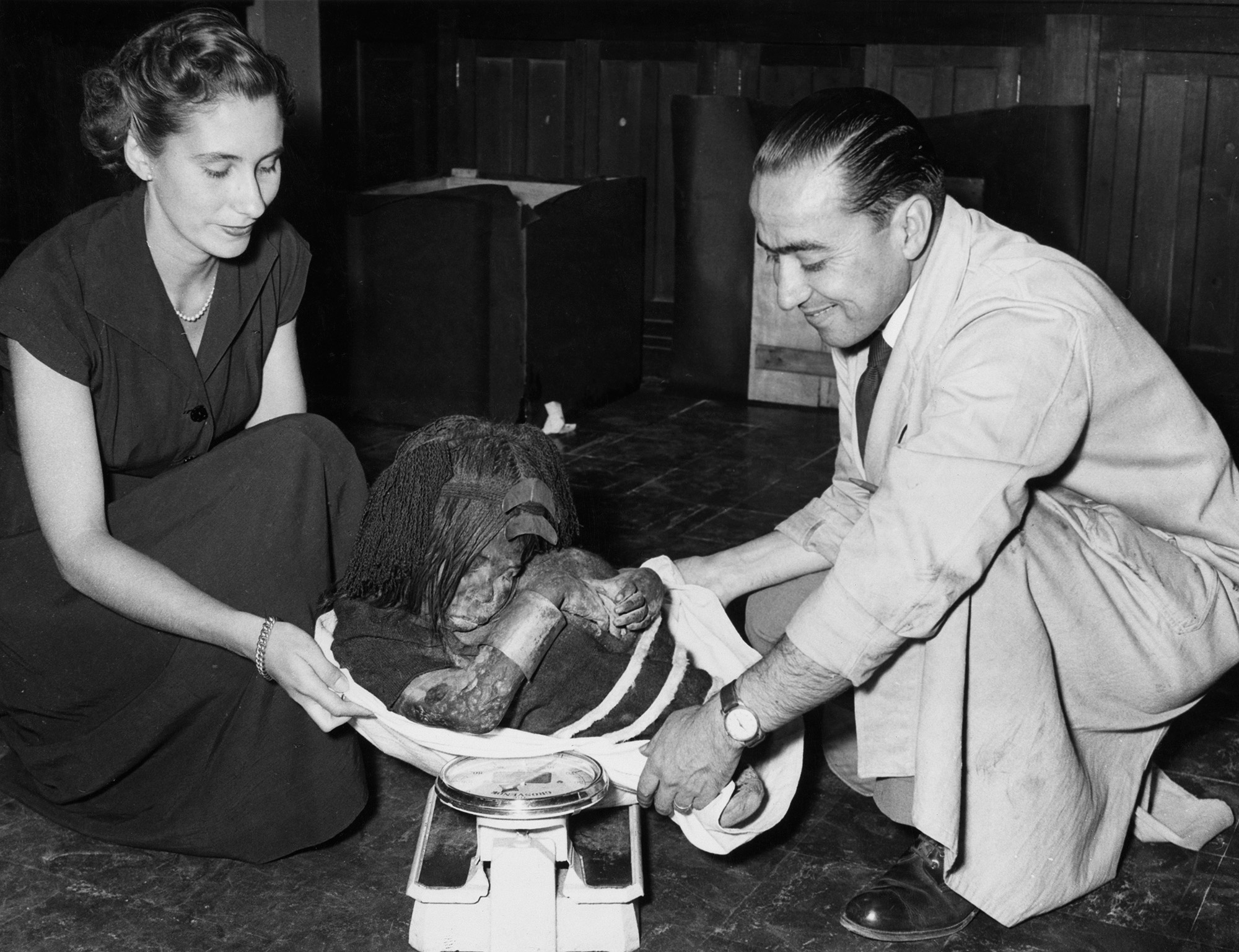 A black and white image shows two people holding a small mummy.