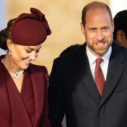 Kate Middleton wears a maroon coat dress and matching hat and smiles while husband Prince William puts his arm around her