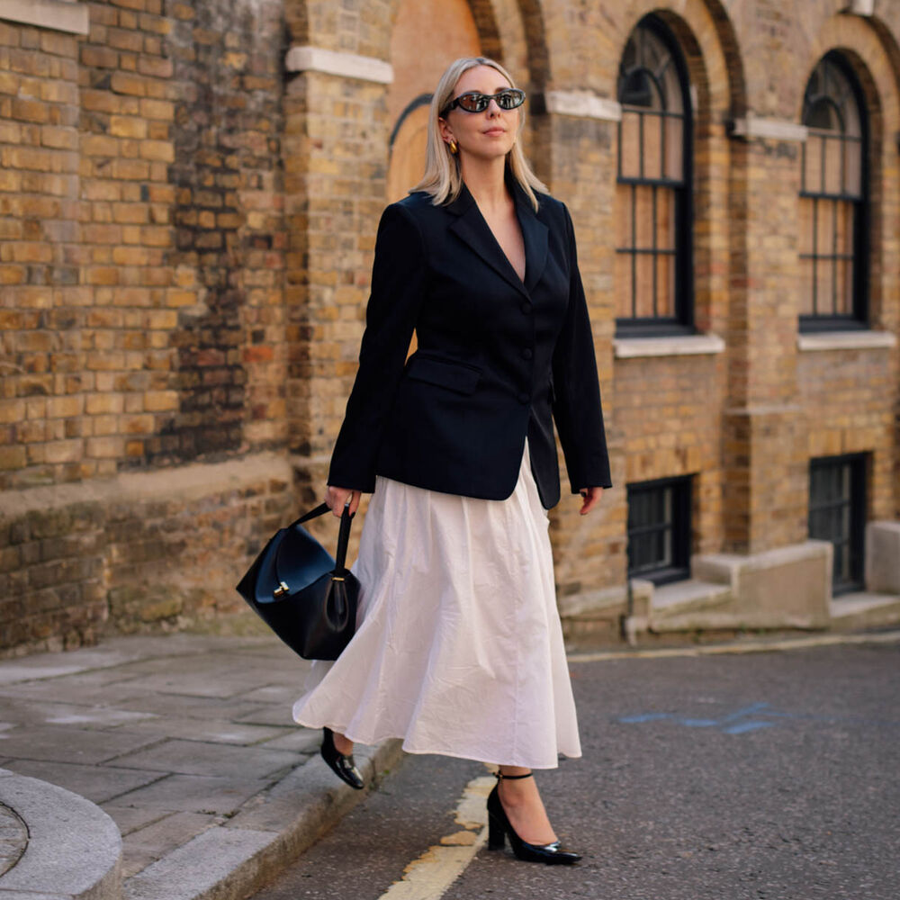 Woman wears oversized blazer, white skirt, and black handbag and heels. 