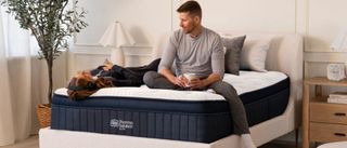 The image shows a white couple with dark hair sitting on a navy blue mattress with a white cover, set within a neutrally decorated bedroom 