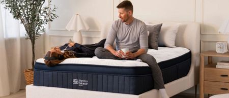 The image shows a white couple with dark hair sitting on a navy blue mattress with a white cover, set within a neutrally decorated bedroom 