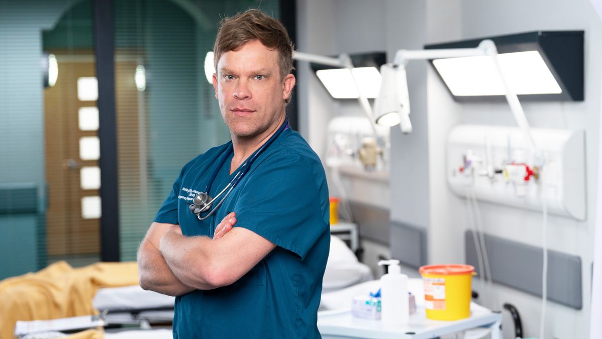 William Beck in a posed shot as Casualty consultant Dylan Keogh in Holby ED, facing the camera looking serious in his blue scrubs with his arms folded. 