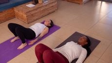 two women lying on their back on exercise mats and tiled floor in a living room setting. with their knees bent and feet flat on the floor. they are both wearing white tops and leggings, one red and one black.