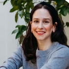 a headshot of professional organizer Dana Reder: a lady with medium length black hair in a long sleeved gray sweater sitting in front of a tall indoor leafy plant looking at the camera smiling
