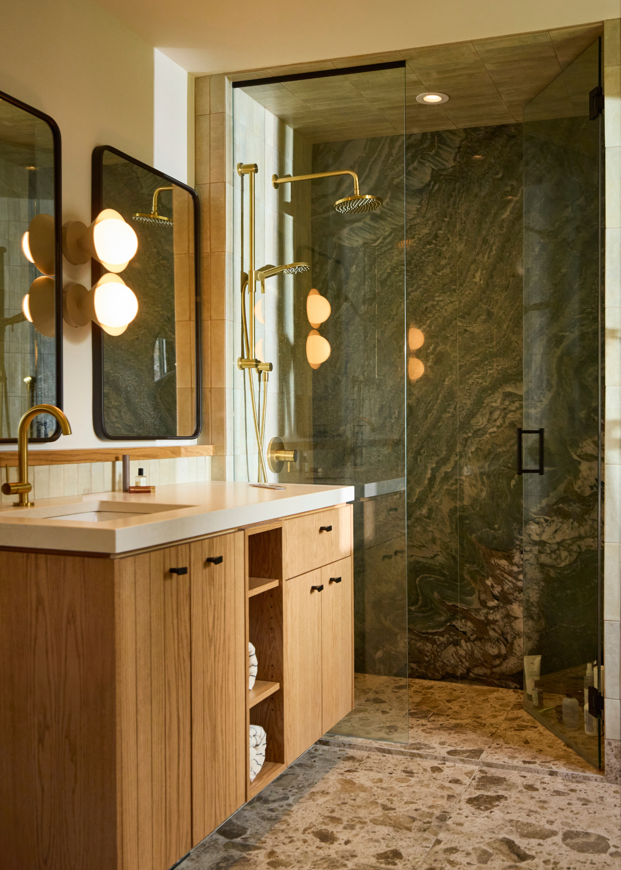 A bathroom with a stone floor, a green marble wall in the shower room, wood bathroom storage with cabinets, a stone counter, and a pair of mirrors with a wall sconce in between