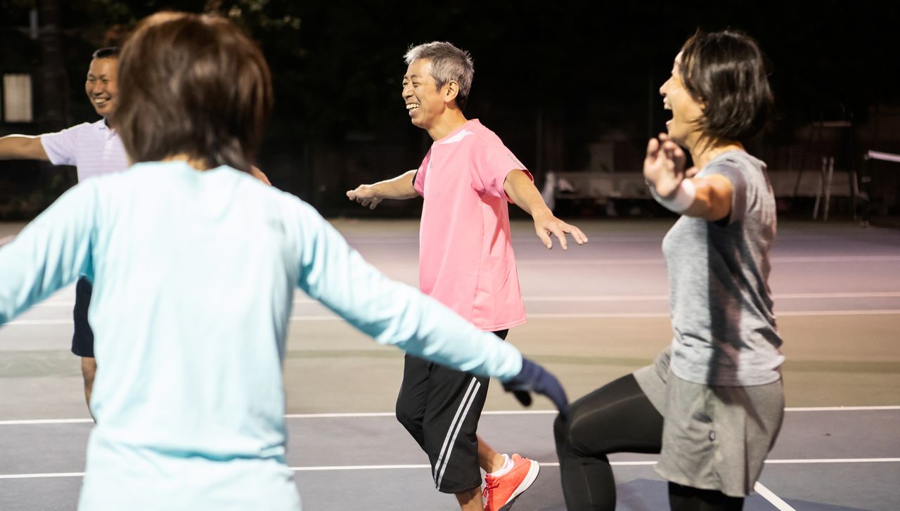 Group of people on a tennis court smile and laugh as they balance on one leg