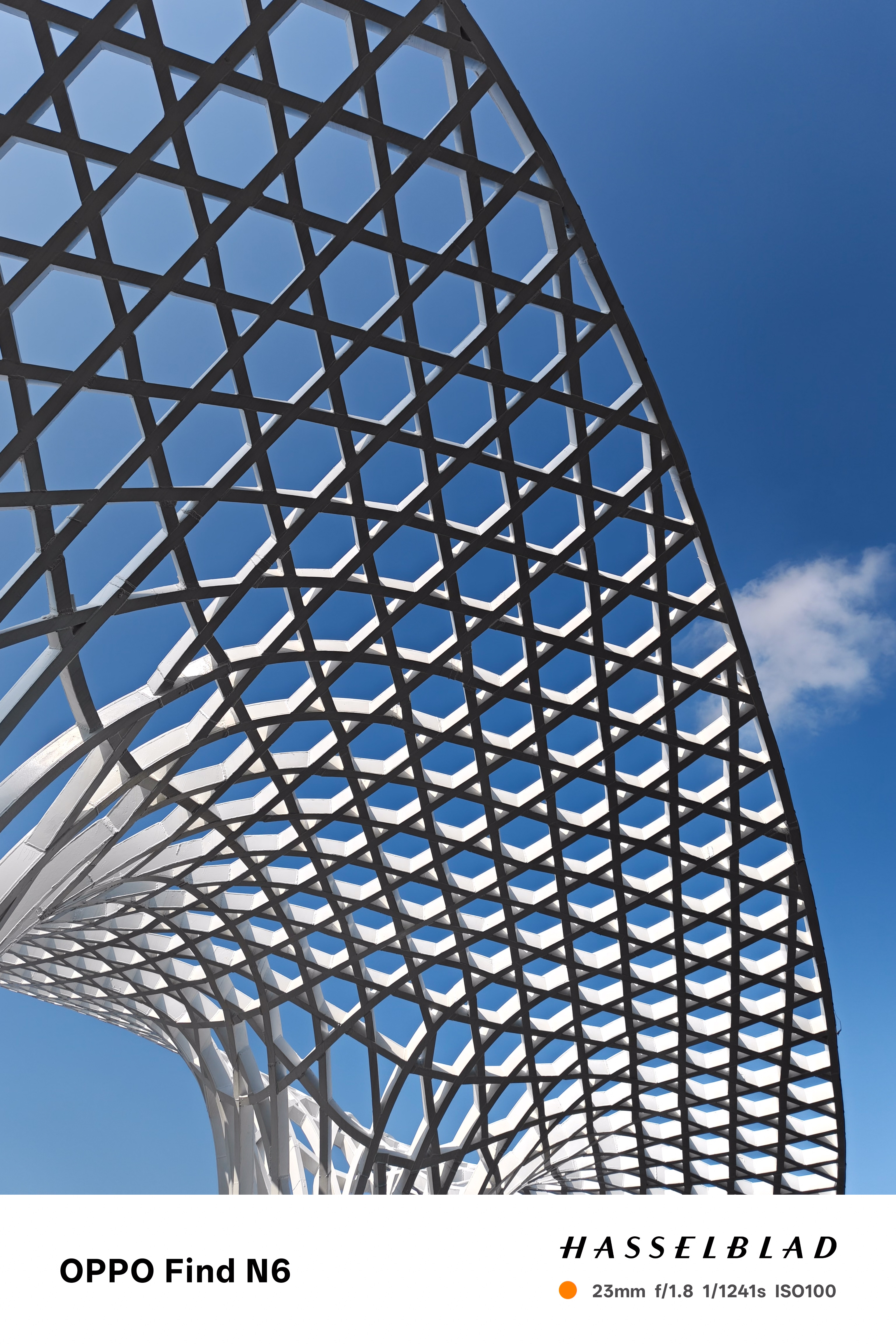 A dramatic low-angle shot of a white architectural pavilion featuring a complex, interlacing honeycomb or lattice roof structure. The white beams form a repeating hexagonal pattern that contrasts sharply with the deep blue sky visible through the gaps.