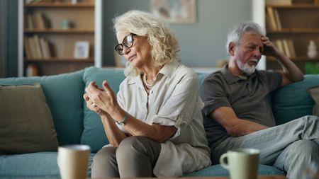 An older couple sit on a sofa together with their backs to each other, clearly not happy.