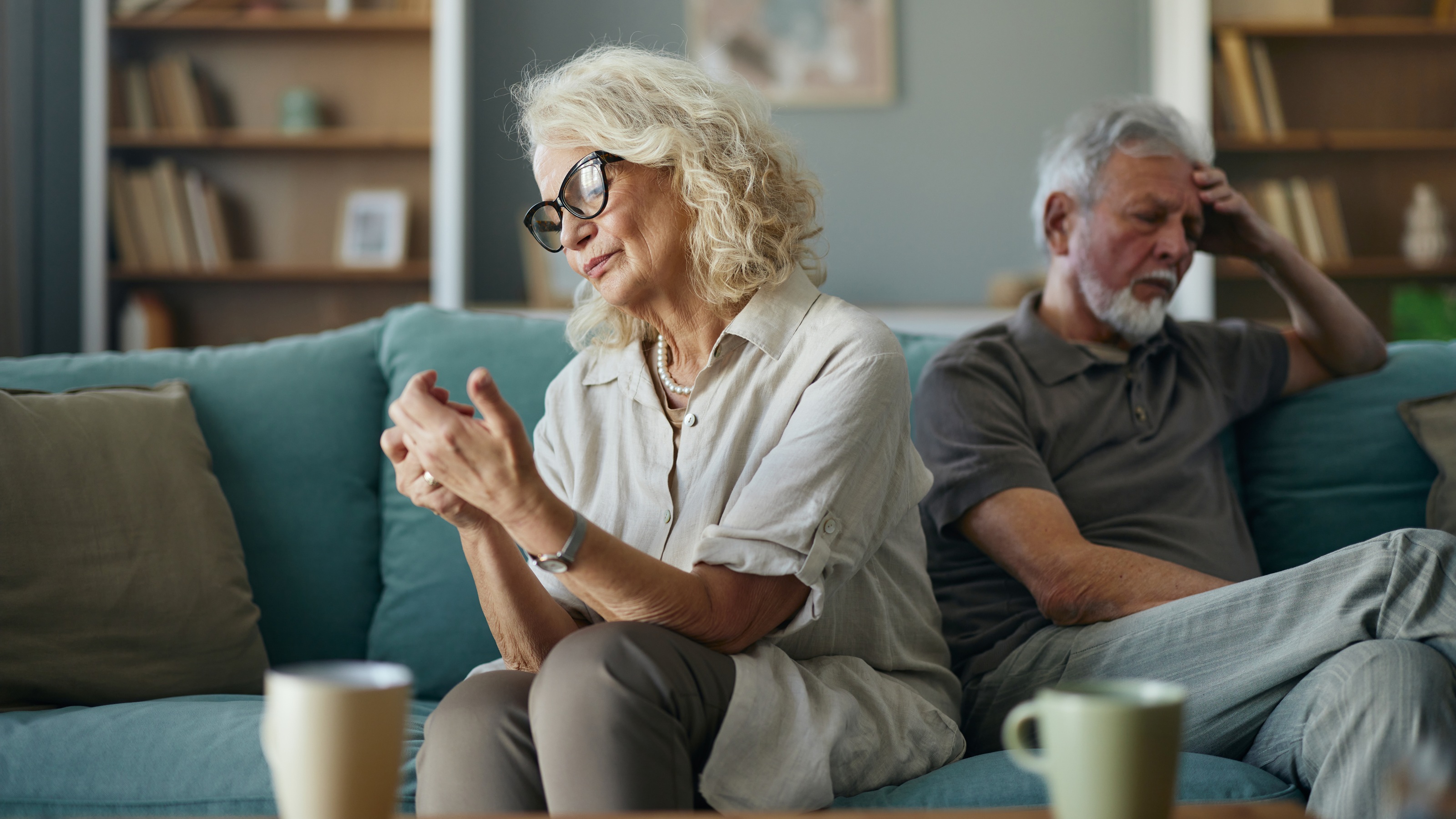 An older couple sit on a sofa together with their backs to each other, clearly not happy.