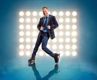 A posed shot of Dancing On Ice's Dan Whiston wearing a black blazer, light blue shirt with a dark blue tie, dark blue trousers and his ice skates, in front of a 9x7 grid of spotlights pointed at the camera