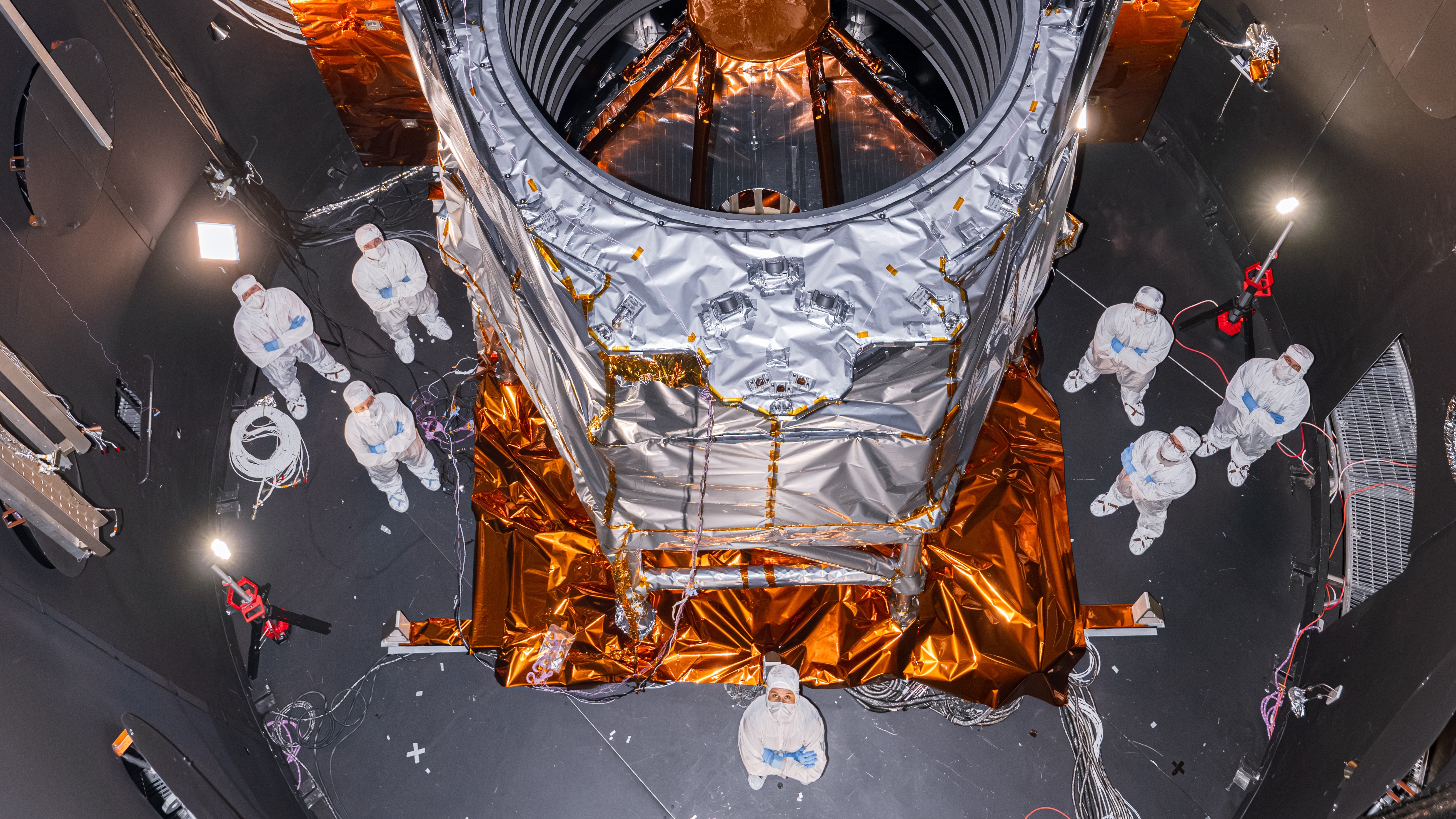 A huge metallic structure in a chamber with lots of people in white clean suits.