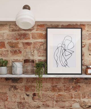 Exposed brick wall with white floating shelf along it with framed print and plant on shelf