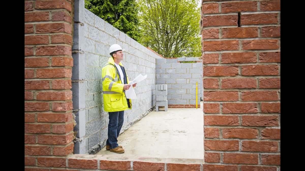 Male building control officer holding building regulations plans and standing within a self build construction site