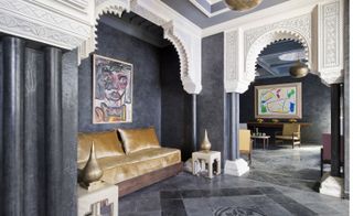 A hotel sitting area with a gold fabric sofa, white side tables with gold ornaments, white arched roof, dark grey pillars, dark grey walls, a desk and square floor tiles in shades of grey.