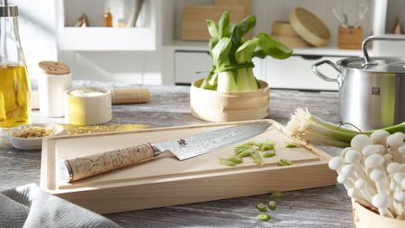 A Japanese knife from ZWILLING on a wood chopping board in a kitchen surrounded by bok choi, mushrooms, spring onions, and olive oil