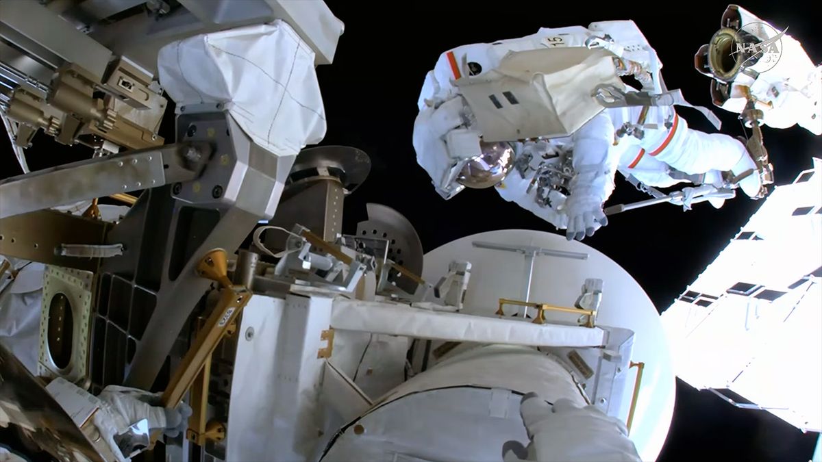 an astronaut in a white spacesuit with red stripes works alongside a large antenna dish outside of a space station