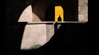 A person stands in a doorway, framed by a vibrant yellow door, creating a striking visual contrast, hard sun creates shadows and light interplay