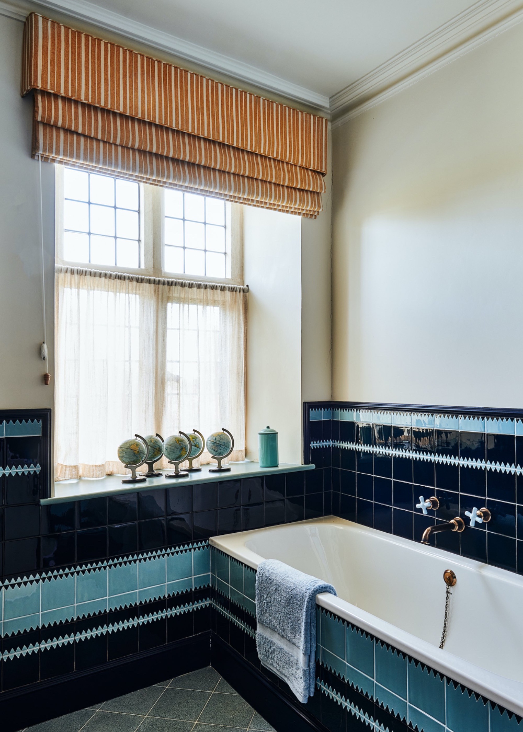 Bathroom with navy and pale blue patterned tiles across the bath panel and lower walls, brass faucets, and an orange striped Roman shade.