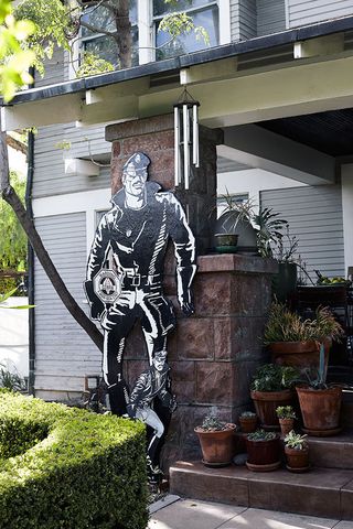 Daytime image at the front entrance of Tom of Finland’s living museum, hedges, stone steps with ceramic pots with plants, brick column with cut out black and white male image, wind chimes hanging down,, stone flagged floor, close up of left wall of house and window, tree and grass shrubs