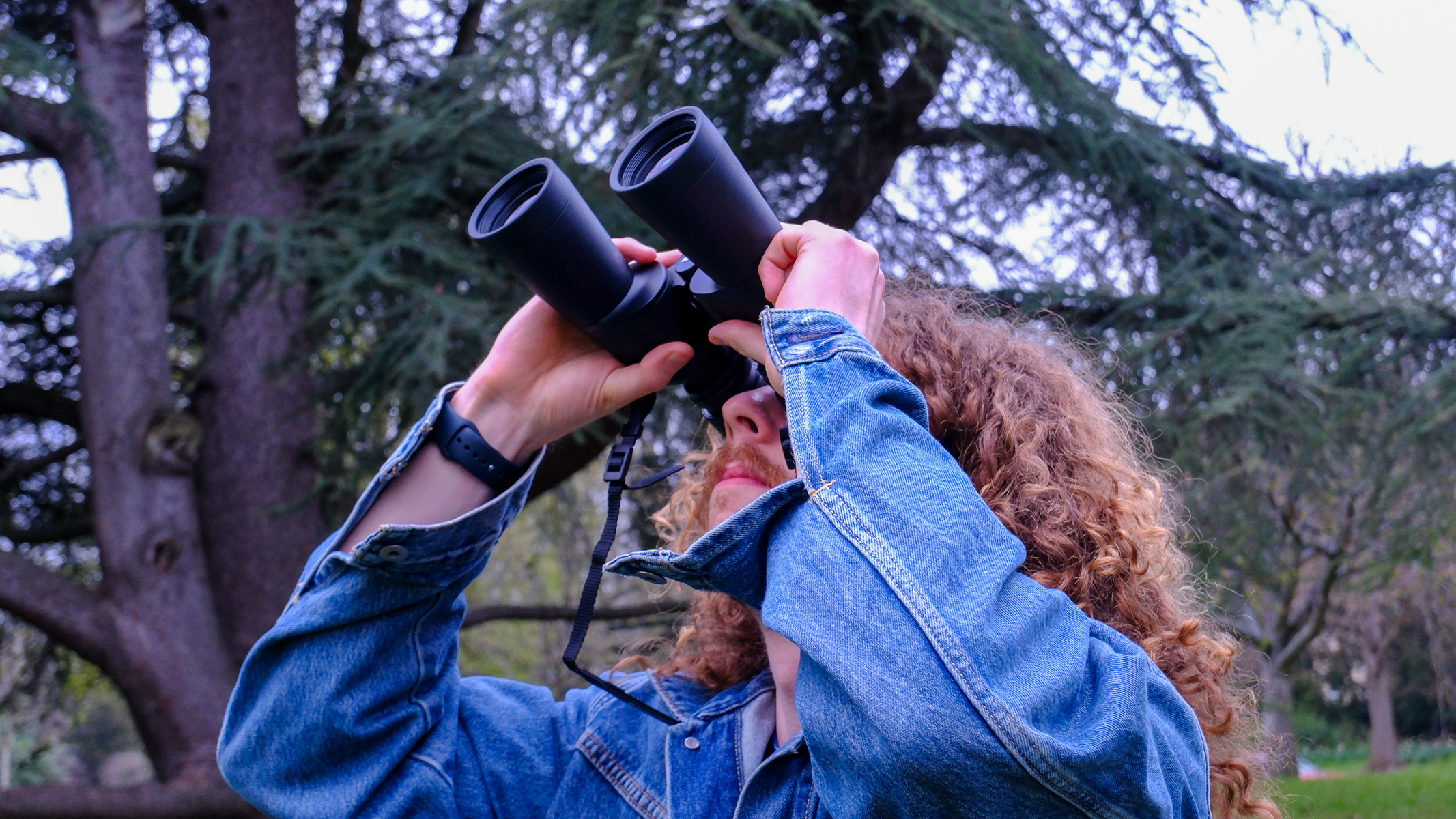 A man with long curly hair looking through the Nikon Action 16x50 binoculars with a tree in the background.