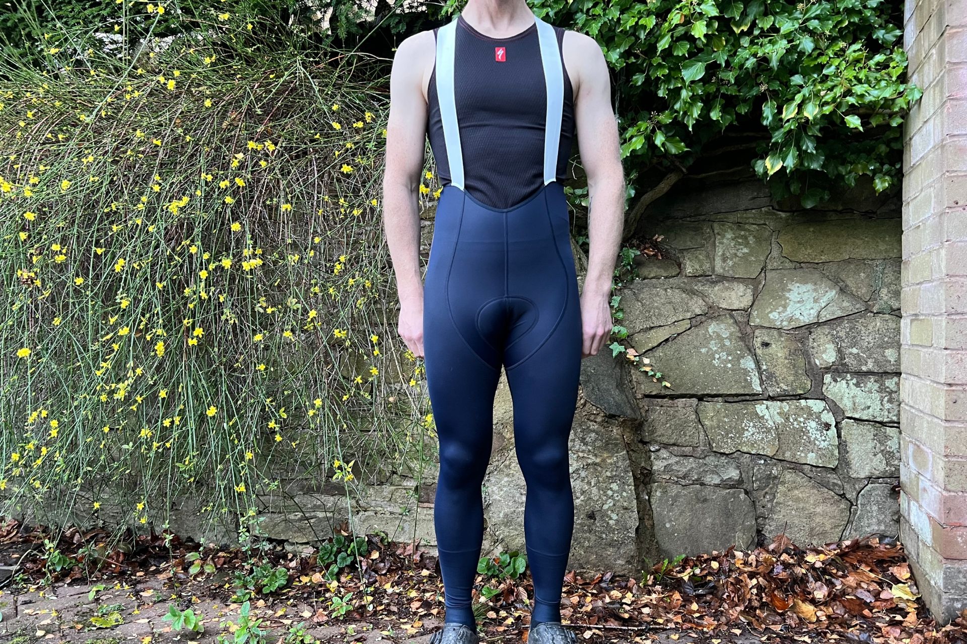 The Rapha Pro Training Tights in navy blue are being worn by a while male who is wearing a sleeveless undervest standing in front of a stone wall with greenery overhanging